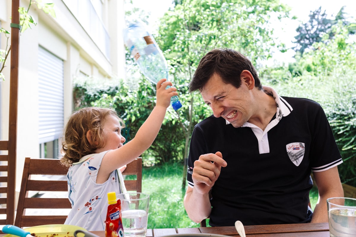 reportage à la maison photo de famille à Rueil Malmaison (92) petite fille tape papa sur la tete avec une bouteille en plastique