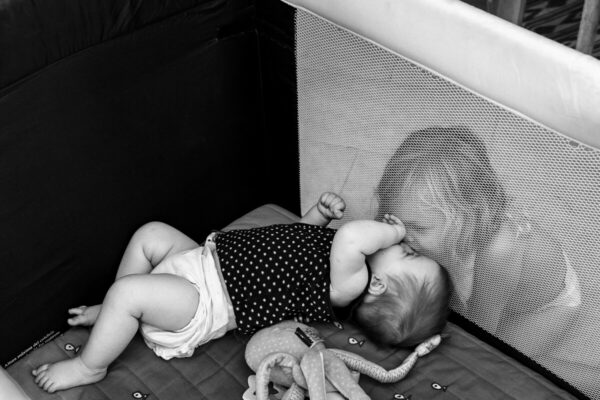 grande soeur qui fait un bisous à sa petite sœur dans un lit parapluie à travers la maille. Reportage de famille à Lille (59)